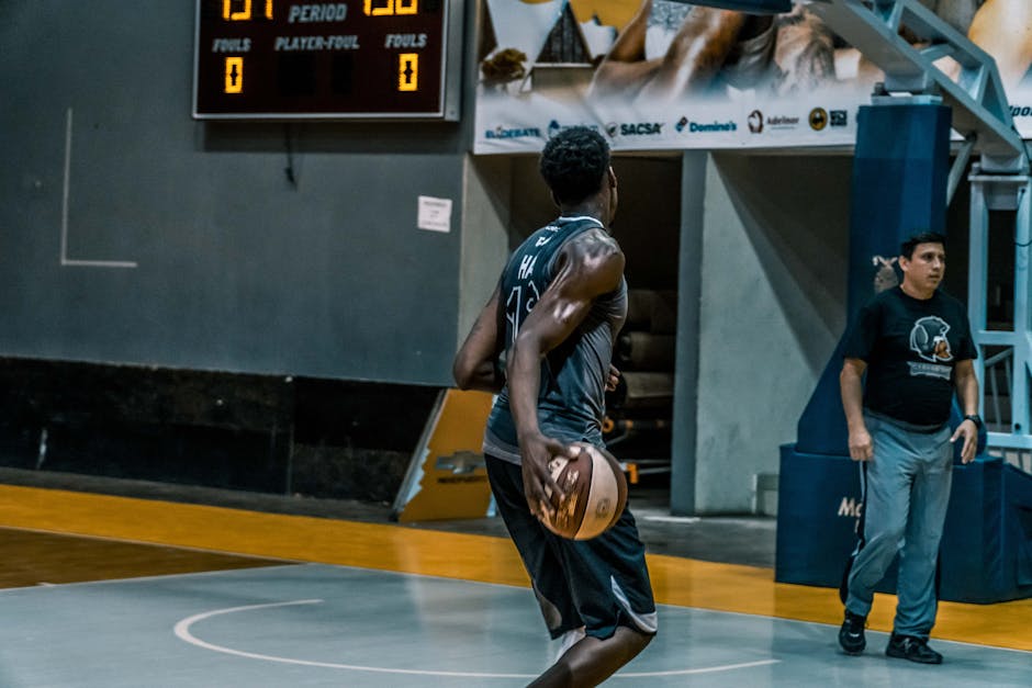 Basketball player performing on an indoor court with scoreboard in background, highlighting athleticism and teamwork.